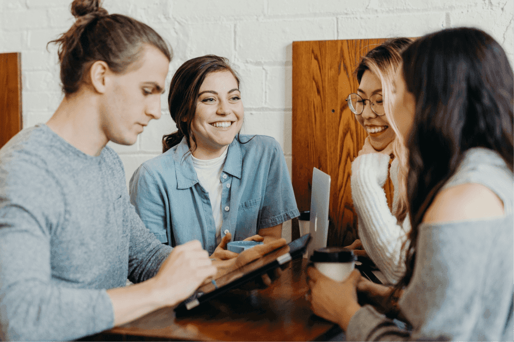 A group of four employees are sat smiling at each other. In between them there is an iPad and several mugs and coffee cups.