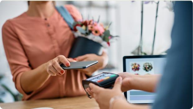 A woman holds a flower pot and is paying a vendor, holding her phone above a card reader. Her face is out of frame.