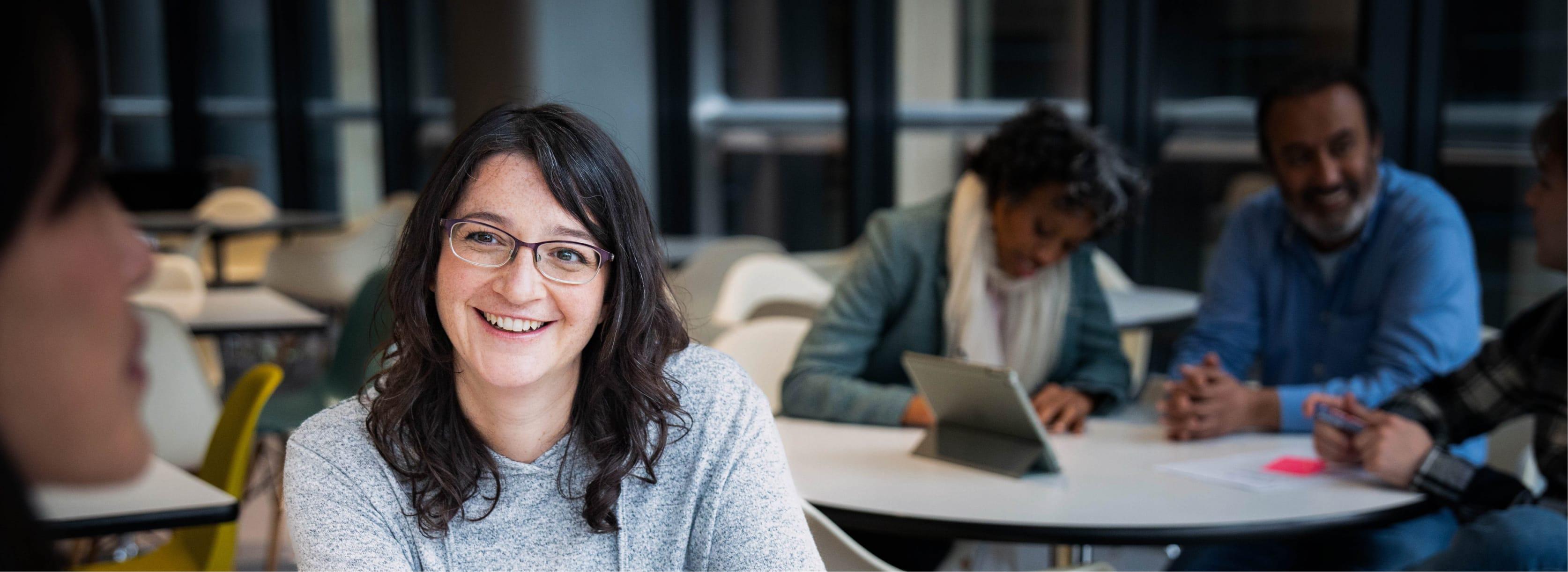 A woman smiles at the camera. Behind her are two people working in the office.