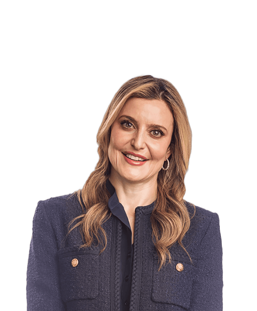 A blonde woman wearing a formal navy jacket smiles into the camera in front of a white blurred