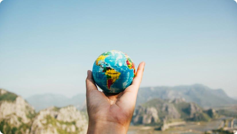 Hand holding a model Earth with the South American continent facing the camera. There are cliffs and a blue sky in the background.