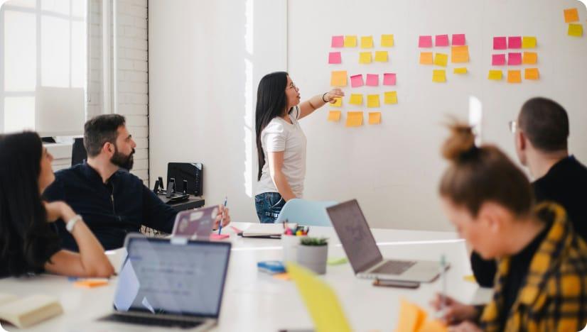 A room full of people with laptops look towards a woman standing in front of a whiteboard, who is pointing towards coloured post-it notes.