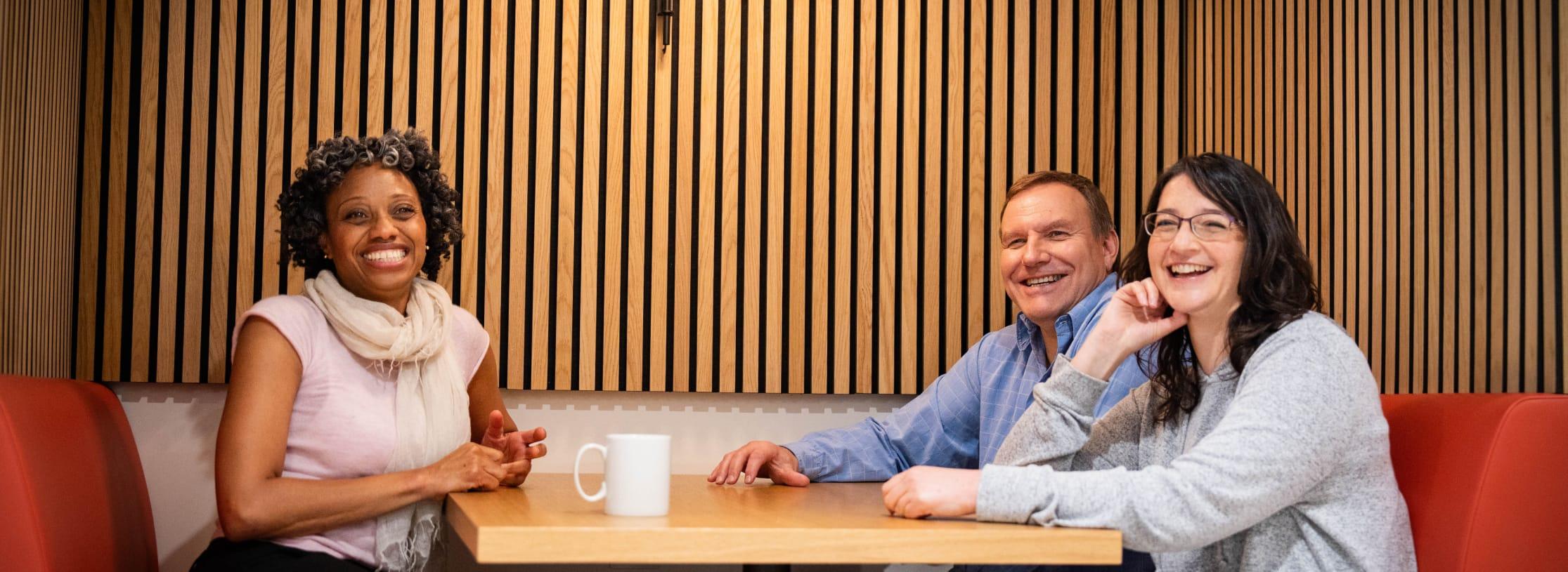 Two women and one man sit at a table booth on red seats and look towards the camera smiling.