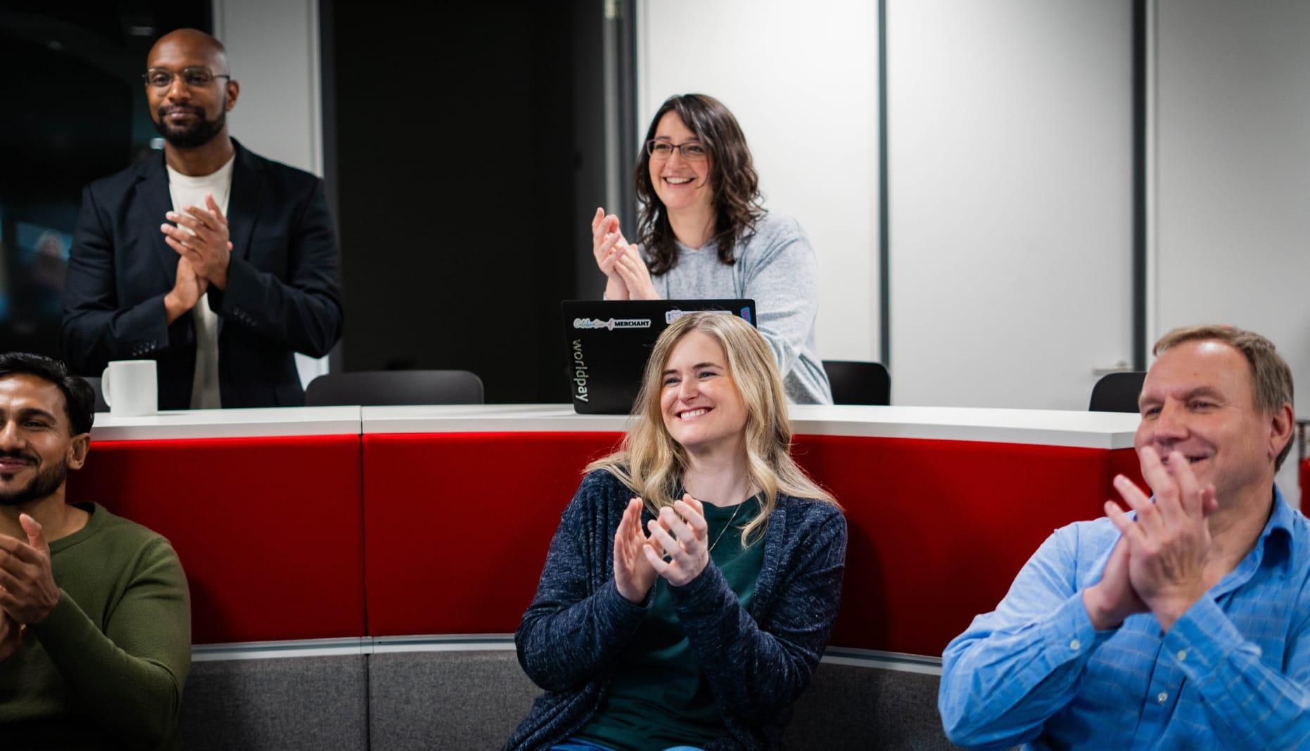 Five people are sitting down on two rows of seats. They are clapping and smiling, and are looking at something out of frame.