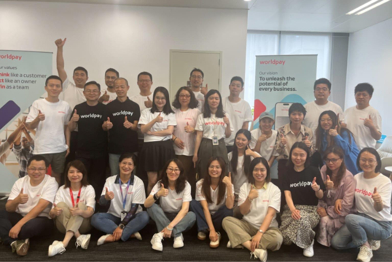 A large group of people wearing Worldpay merch stand together in front of banners with the Worldpay values and vision. They all have their thumbs up.