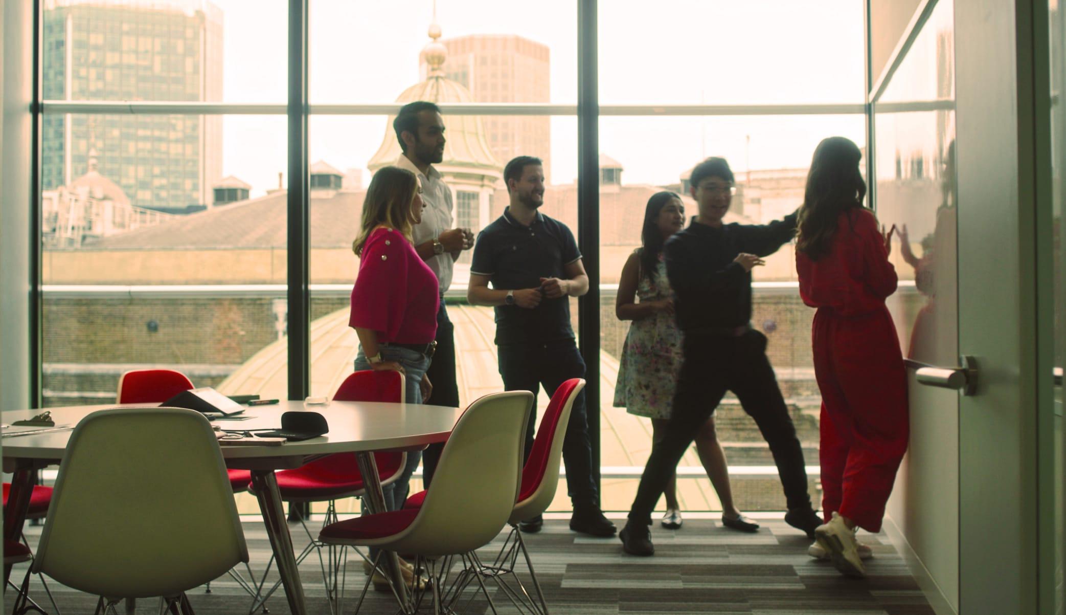 Multiple people, backlit by a large window, stand behind a circular table in an office. They’re looking at something out of frame.