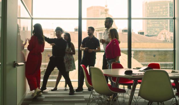 Multiple people, backlit by a large window, stand behind a circular table in an office. They’re looking at something out of frame.