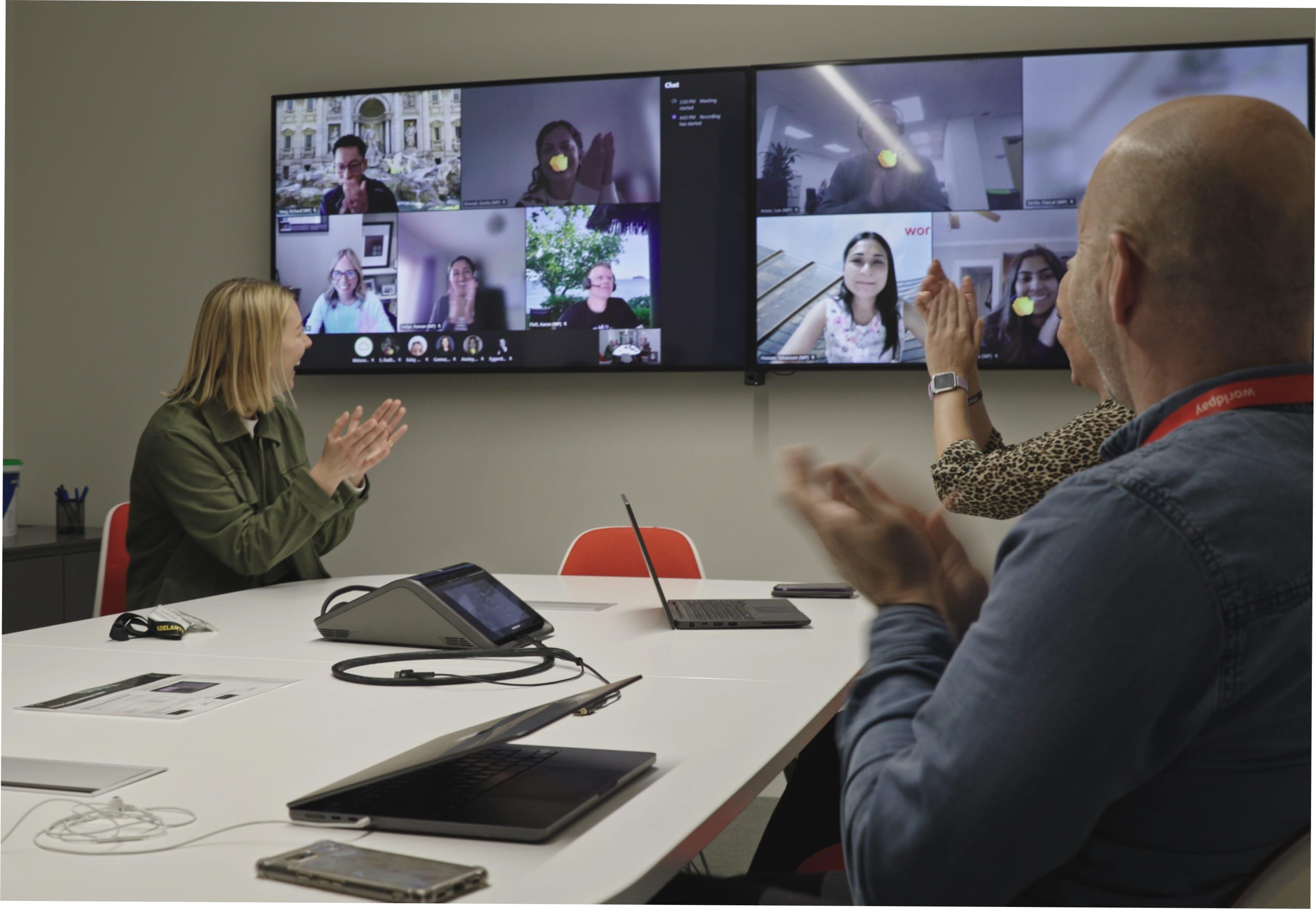 A woman claps whilst laughing towards a TV screen in a meeting room that has multiple members filling the screen