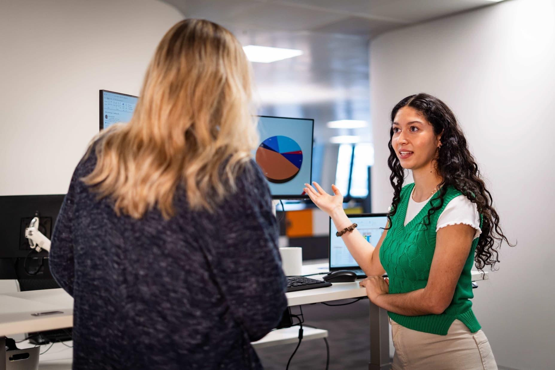 Two females talking in front of screen with pie chart on, one pointing at the screen.