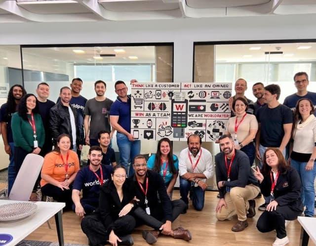 About 20 diverse team members posing in a modern office around a banner reading “Think like a customer, Act like an owner, Win as a team, We are Worldpayer” with black-and-white icons and a red “W” logo.