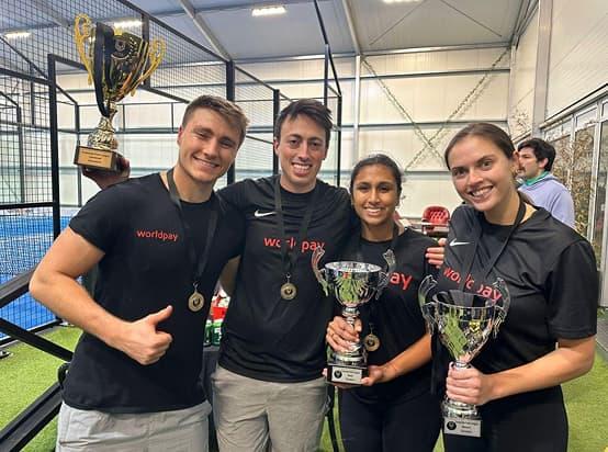 Four smiling teammates in black “Worldpay” shirts pose indoors holding trophies and medals, celebrating a sports victory on artificial turf next to a large gold cup on a pedestal.