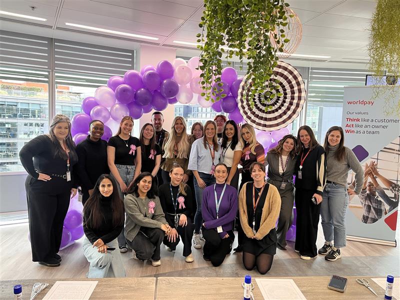 Group of people posing in an office with purple and white balloon arch, under hanging plants. A Worldpay banner with company values is visible on the right.